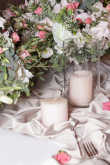 A stylish wedding decor of dishes, white bouquets with flowers, candles and glasses stand on a white tablecloth of a served table in a restaurant. Side view, selective focus