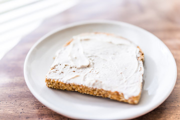 Closeup of slice piece of whole wheat sprouted toasted grain bread on plate with white cream cheese spread on table macro