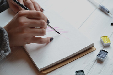 Woman paints in a sketchbook. Tools for painting on the white wooden desk. Painter work.