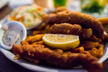 Macro closeup of deep fried fish and chips battered filets brown golden crust in restaurant with lemon slice, tartar sauce, side dish on table in evening restaurant dinner