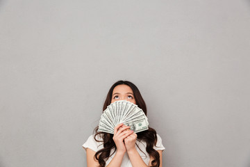 Photo of asian rich woman 20s covering face with fan of money dollar banknotes and looking upward, isolated over gray background