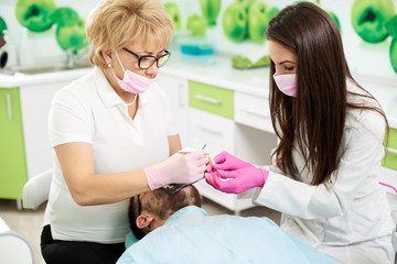 Obraz na płótnie Canvas Female dentist specialist and her young brunette assistant are giving dental treatment to a male client of a modern dentistry.