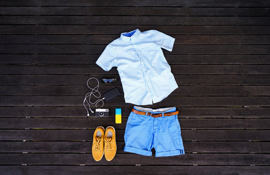 Man summer clothes collage flat lay isolated on dark wood background. Summer outfit of casual man desk top view fashion accessories: shirt, shorts, sunglasses, camera, smartphone. Holiday travel