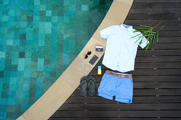 Man summer clothes collage flat lay isolated on dark wood background with swimming pool. Summer outfit of casual man desk top view fashion accessories: shirt, shorts, sunglasses, camera, smartphone