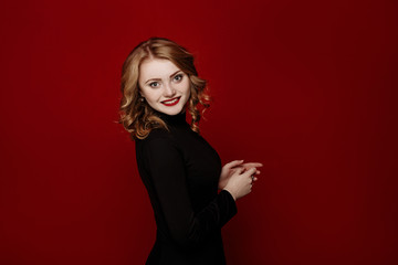 young cheerful red-haired girl in black clothes posing in the studio isolated on red background. body language and emotions. woman smiling into camera rejoicing her success. 