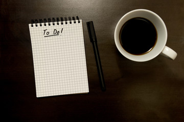 Fototapeta na wymiar To do list on gridded spiral notepad with pen and cup of coffee - from above on dark timber desktop top background - with copy space
