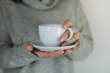 Young woman in grey cozy pullover holding white cup in hands. White mug for man, woman, gift. Mockup for designs.