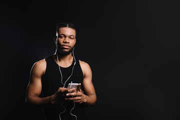 young african american sportsman in earphones holding smartphone and looking at camera isolated on black