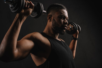 Fototapeta na wymiar concentrated muscular african american man exercising with dumbbells isolated on black