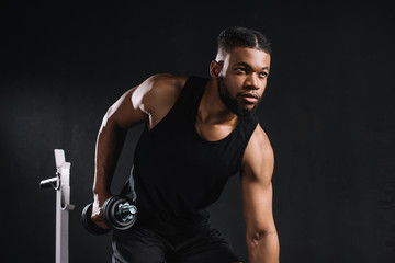 Fototapeta na wymiar young african american sportsman exercising with dumbbell and looking away on black