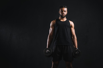 Fototapeta na wymiar young muscular african american sportsman holding dumbbells and looking at camera isolated on black