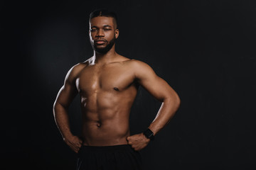 Fototapeta na wymiar muscular young african american sportsman standing with hands on waist and looking at camera isolated on black