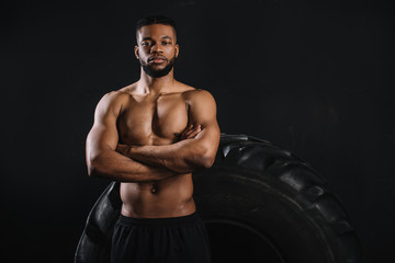 Fototapeta na wymiar muscular shirtless african american sportsman standing with crossed arms near big tyre and looking at camera isolated on black