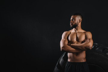 Fototapeta na wymiar muscular shirtless african american sportsman standing with crossed arms near big tyre and looking away isolated on black