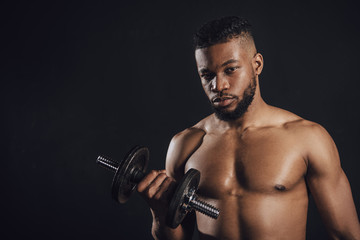 Fototapeta na wymiar muscular shirtless african american sportsman training with dumbbell and looking at camera isolated on black