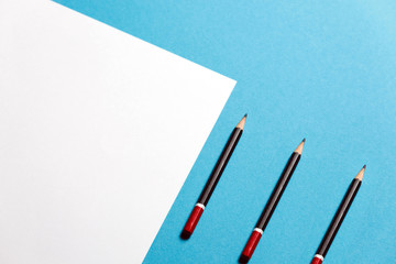 Three Pencils Lies On A Paper Sheet On Blue Background, Top View. Office Space Creativity Minimalism Concept