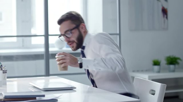 Young handsome entrepreneur walking into office, drinking coffee, sitting at desk and turning on laptop