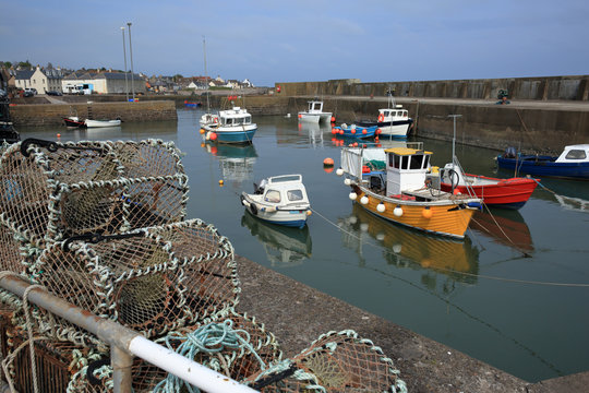 The still tranquil harbour at Johnshaven in north east Scotland UK. Small yellow red and green fishing boats float in a harbour surrounded by a concrete sea wall. Lobster pots stored by fishing boats