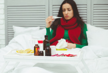 Portrait of a young sick woman in a red scarf measures the temperature with a thermometer in the bed at home. The concept of health and disease.