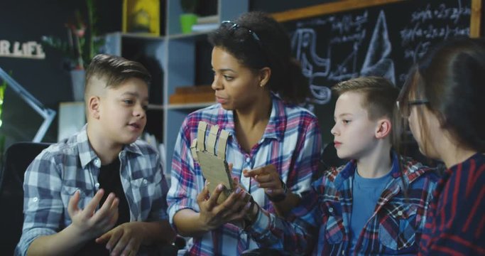 Young woman with class of children talking about 3d printed prosthesis of hand made of cardboard.