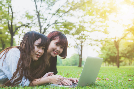 Young asian women lying on grass and using laptop and typing. Girls hands on keyboard. Distance learning concept. Happy hipster young asian women working on laptop in park. Student studying outdoors.