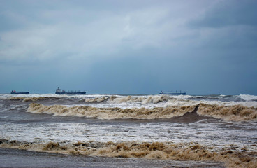 The incoming large waves on the shore and the ship on the horizon in the background of a cloudy sky.