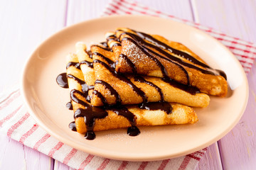 Crepes with chocolate syrup on purple wooden background.