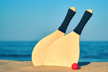 Set for a game of beach tennis on the background of sea and mountains close-up. Wooden rackets and red ball in the sand                             