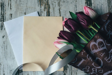 a bouquet of white and pink and purple tulips in a paper purple wrapper on a wooden white board with paper envelopes and a gray ribbon