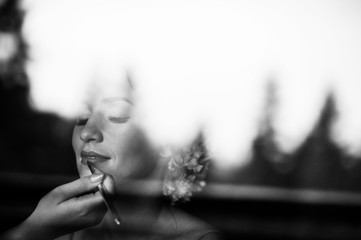 Obraz premium Black and white photo of bride through the window glass, the makeup artist put on lipstick on her lips