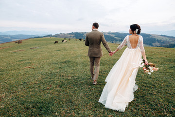 Fototapeta na wymiar The groom and bride in beige dress with naked back are walking on green hill with mountains on background