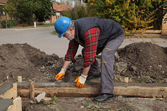 Worker spreading concrete in formwork for wall foundation using trowel, real people working