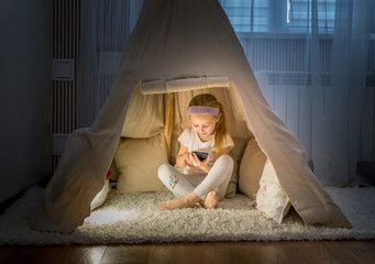 Little girl with smartphone sitting in teepee tent