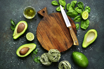 Fototapeta na wymiar Uncooked spinach pasta (tagliatelle) and green vegetables (spinach, lime, avocado) with empty wooden cutting board.Top view with copy space .