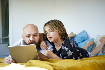 Father showing tablet computer to his son while lying on bed at home