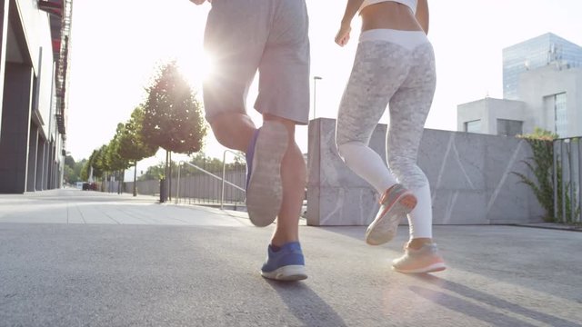 SLOW MOTION, LOW ANGLE, LENS FLARE: Fit Caucasian couple bonding by running along the asphalt city streets on a sunny spring morning. Two fit friends exercising outdoors on a beautiful spring morning.