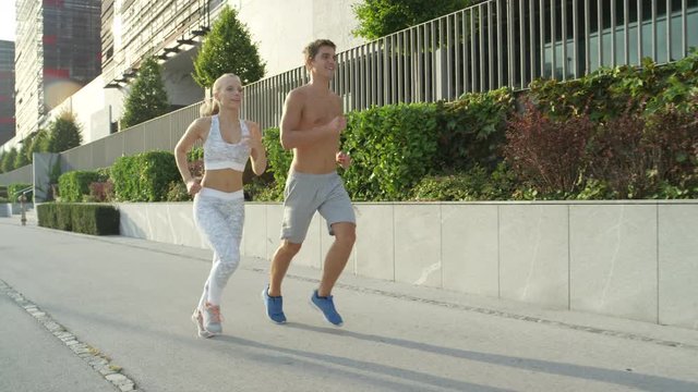 SLOW MOTION: Active girl and boy on a delightful jog in sunny spring in big city. Fit couple enjoy spending time together jogging through sunlit city streets in summer. Young athletic people running
