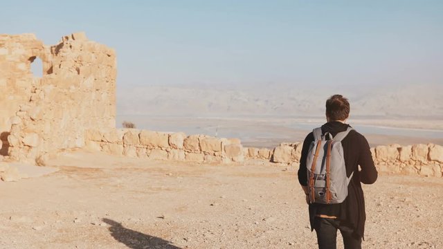 Man with backpack walks on desert mountain top. Casual journalist blogger takes photos of Dead Sea scenery. Israel 4K.