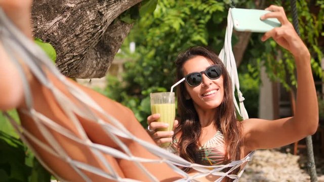 Mixed Race Young Tourist Woman Taking Smartphone Selfie Picture Relaxing in Hammock on Tropical Island. Girl Using Mobile Phone at the Beach. 4K, Slowmotion. Nusa Penida, Bali, Indonesia.