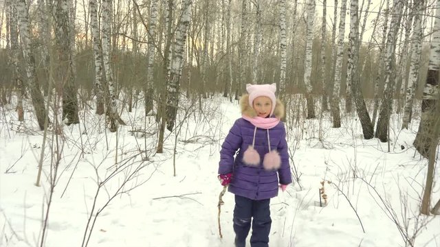 Little girl runs through birch woods with a stick. Young girl happy jokes and falls to snow in winter forest.