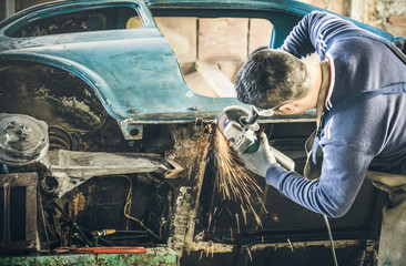 Young man mechanical worker repairing old vintage car body with electric grider in messy garage - Work safety with protection wear - Niche expertise concept with guy working with technical equipment