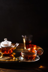 Small glass teapot and glasses with hot black tea, dried rose petals, pocket magnifier on golden chain, squeezed orange slice on golden tray. Evening light. Side view.
