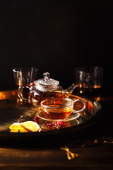 Small glass teapot and glasses with hot black tea, dried rose petals, pocket magnifier on golden chain, squeezed orange slice on golden tray. Evening light. Dark background