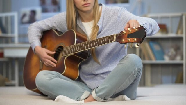 Female musician tuning the guitar on home sofa, enjoying melody, practicing