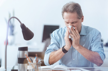 Cold symptoms. Cheerless depressed adult man sitting at the table and suffering from the running...