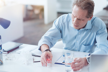 My medicine. Nice handsome pleasant man holding a glass of water and taking medicine while sitting in his office