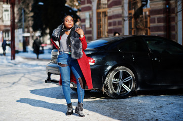 Rich african american girl in red coat and fur on winter street at sunny weather against black business car. Black stylish woman.