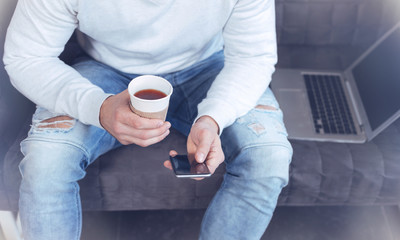 Waiting for email. Delighted man holding cup with tea and playing with his telephone while sitting on the sofa