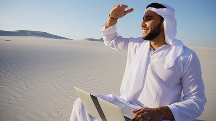 Stately Arabian UAE Sheikh entrepreneur holds in hands and examines project on paper, compares with future site for construction and transfers data to laptop, sitting on white sand in bottomless wide