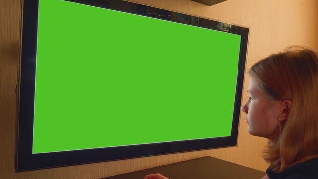 Girl sits in front of TV set with green screen and watches it.